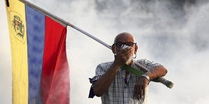 Ein Mann mit venezolanischer Flagge hält sich ein Tuch gegen Gasschwaden vor das Gesicht