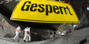 Besucher in Schutzanzügen laufen durch einen dunklen Schacht. Im Vordergrund ein Schild: "Gesperrt"