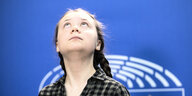Greta Thunberg steht vor einer blauen Wand und blickt bedeutungsschwanger nach oben