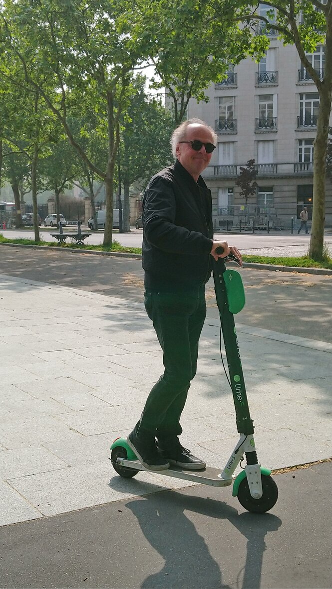 taz-Frankreich-Korrespondent, ein älterer Mann mit weißem Haar, steht auf einem Elektroroller