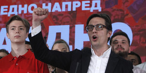 Ein Mann mit schwarzer Brille streckt auf einer Bühne mit rotem Hintergrund die rechte Faust in die Höhe.