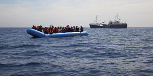 Ein Schlaucboot mit vielen Menschen, im Hintergrund nähert sich ein größeres Schiff