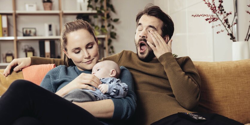 Ein Vater reibt sich gähnend die Augen, während die Mutter das Baby auf dem Arm hält.