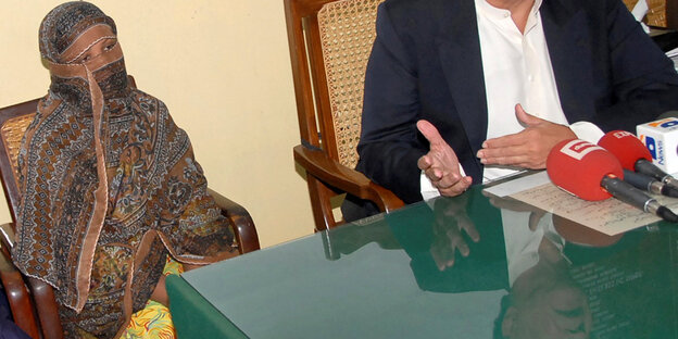 Asia Bibi sitzt verschleiert an einem Tisch, neben ihr die Hände von einem Mann, der in ein Mikro spricht