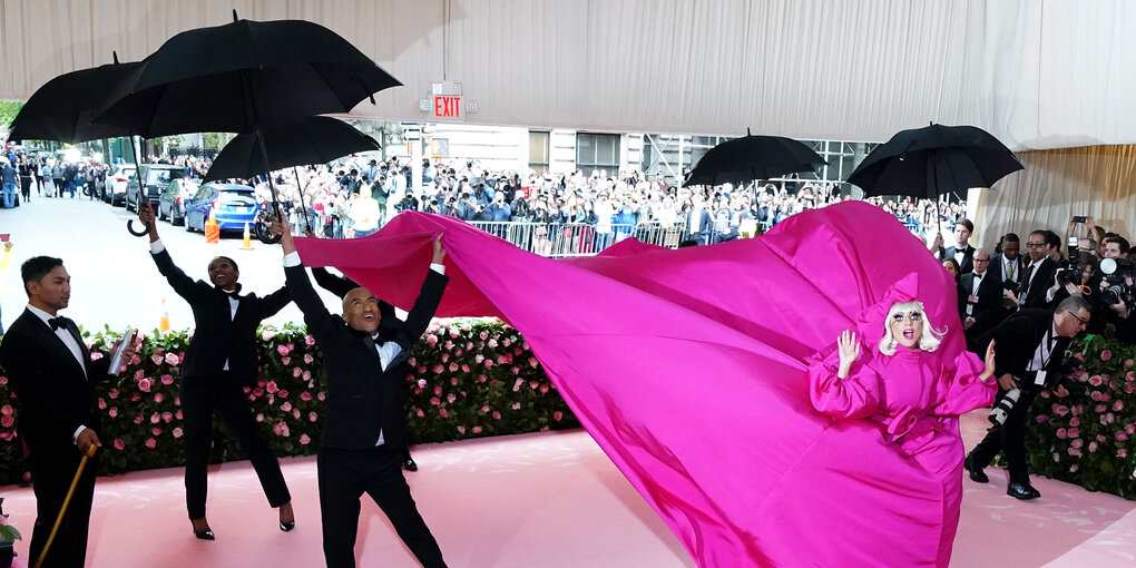 Lady Gaga bei der Met Gala im meterlangen pinken Kleid umringt von schwarzgekleideten Männern mit Regenschirmen.