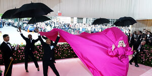 Lady Gaga bei der Met Gala im meterlangen pinken Kleid umringt von schwarzgekleideten Männern mit Regenschirmen.