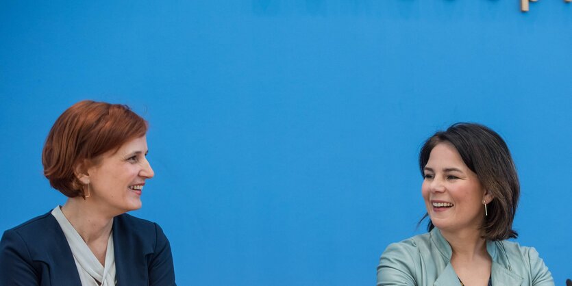 Katja Kipping und Annalena Baerbock strahlen einander an, sie sitzen vor einer blauen Wand