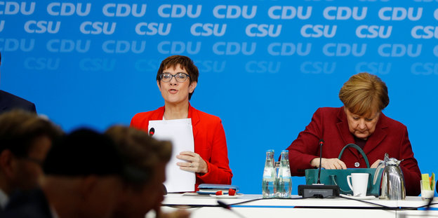 Annegret Kramp-Karrenbauer und Angela Merkel sitzen auf einem Podium nebeneinander. Kanzlerin Merkel sucht etwas in ihrer Handtasche