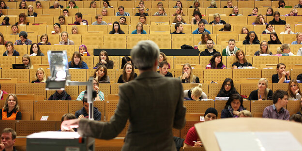 Eine Professorin hält eine Vorlesung in der Universität, vor ihr sitzen mehrere Studierende in einem Saal.