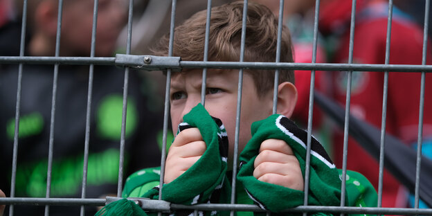 Ein kleiner Junge mit dem grünen Fan-Schal Hannovers steht hinter einem Zaun.