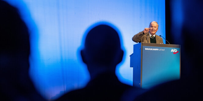 Alexander Gauland vor einer blauen Wand am Rednerpult, im Vordergrund die dunklen Schatten zweier Köpfe