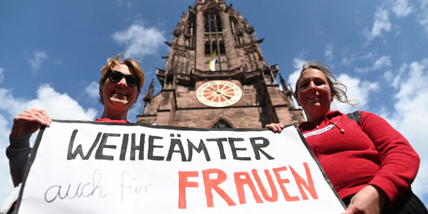 Zwei Frauen stehen vor dem Freiburger Münster und halten ein Banner mit der Aufschrift "Weiheämter auch für Frauen" in die Höhe