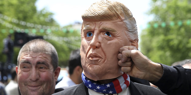 Ein Mann steht neben einer Trump-Maske, diese wird von einem anderen Mann geschlagen