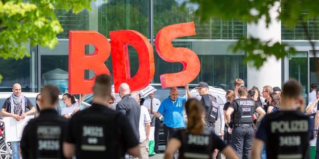 AktivistInnen halten riesige Buchstaben "BDS" hoch, davor stehen PolizistInnen