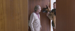 Zwei Männer stehen sich an einer Tür gegenüber. Einer hält einen offenbar ausgestopten Hund im Arm