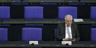 Bundesinnenminister Horst Seehofer sitzt im Bundestag und tippt auf seinem Handy, die blauen Stühle um ihn herum sind leer