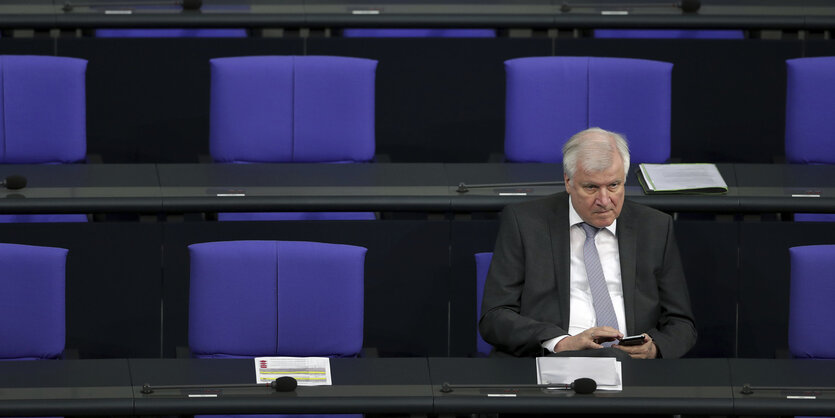 Bundesinnenminister Horst Seehofer sitzt im Bundestag und tippt auf seinem Handy, die blauen Stühle um ihn herum sind leer