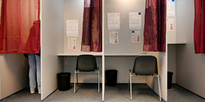 links ein Wähler hinter einem Vorhang, rechts drei leere Wahlkabinen