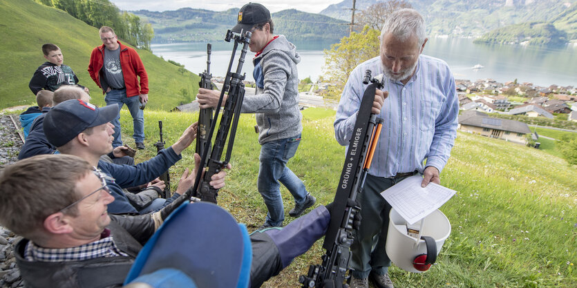 Zwei Männer inspizieren ihre Waffen, im Hintergrund ein Bergpanorama und ein See