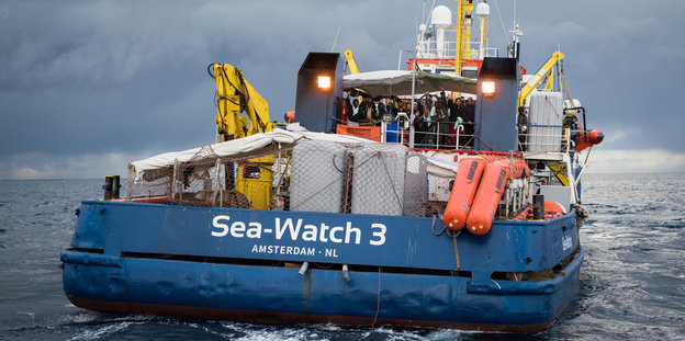 Auf dem Seenotrettungsschiff „Sea Watch 3“ stehen Gerettete und Crewmitglieder und winken.
