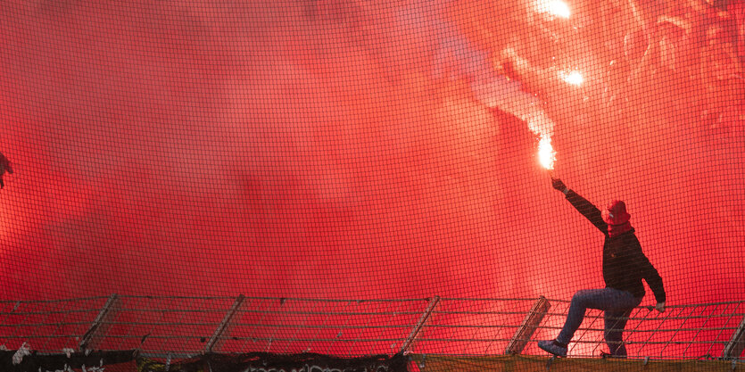 Ein Mann sitzt auf einem Stadionzaun und schwenkt eine bengalische Fackel