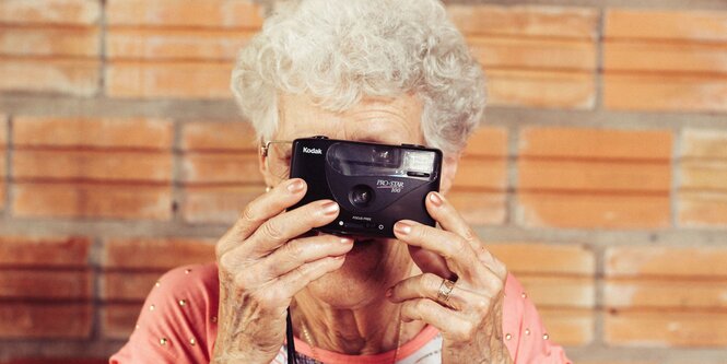 Eine ältere Frau mit weißem Haar und rosa Shirt guckt durch einen Fotoapparat