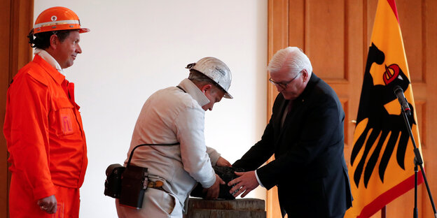 Bundespräsident Steinmeier und ein Bauarbeiter fassen zusammen einen schweren Stein an - im Hintergrund eine Deutschlandfahne