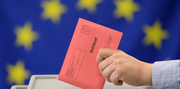 Eine Person wirft einen Umschlag in eine Wahlurne, dahinter eine Fahne der Europäischen Union