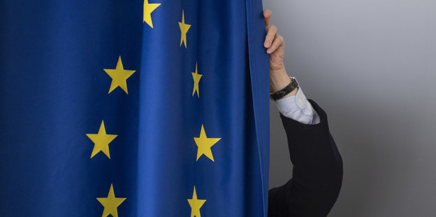 Ein Mann in Anzug steht hinter einer sehr großen EU-Flagge und zieht sie wie einen Vorhang.
