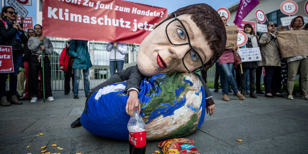 Eine Aktivistin von Campact hat eine Großmaske auf, die die CDU-Vorsitzende zeigt und liegt neben einer offenen Chipstüte und Softgetränk auf einem Sitzsack, der mit einem Erdkugel-Motiv bedruckt ist.