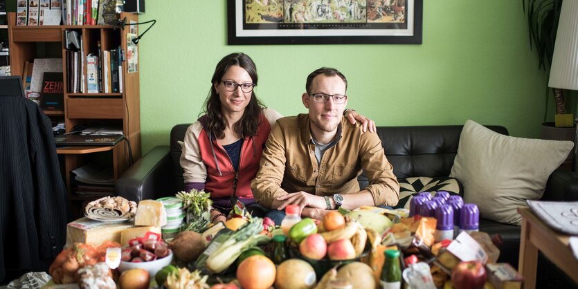 Eine jung Frau und ein junger Mann sitzen auf dem Sofa. Vor ihnen auf dem Tisch ist eine Fülle von lebensmtiteln ausgebreitet