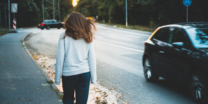 Eine junge Frau, die nur von hinten zu sehen ist, läuft auf einem Bürgersteig entlang. Ein Auto fährt auf der Straße daneben in die entgegengesetzte Richtung.