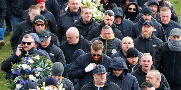 Mehrere schwarztragende Männer, viele davon glatzköpfig, tragen einen Sarg mit Blumen