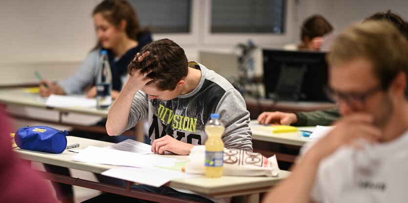 Ein Schüler sitzt an seinem Platz und liest seine Aufgaben, er fasst sich mit der rechten Hand in die Haare.