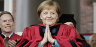 Angela Merkel trägt einen rot-schwarzen Talar und faltet ihre Hände