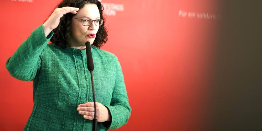 Andrea Nahles steht vor einem Mikro und hält die rechte Hand in Kopfhöhe