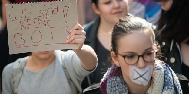 Demoteilnehmerin mit zugeklebtem Mund, hinter ihr ein Schild auf dem steht: "Wir sind keine Bots"