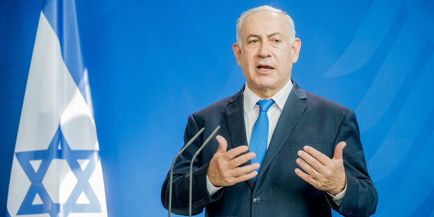 Netanjahu steht vor einer Israelfahne und redet in ein Mikro
