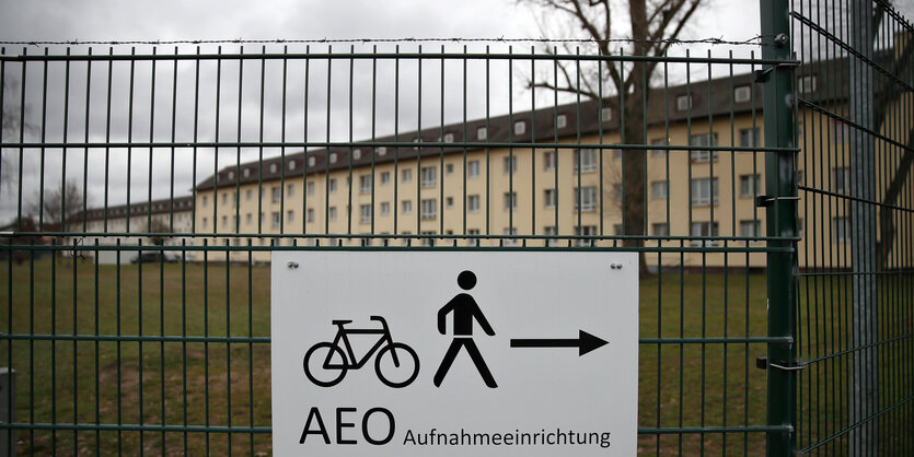 Hinter einem Zaun mit Stacheldraht ist ein Gebäude zu sehen, auf einem Schild steht "Aufnahmeeinrichtung"