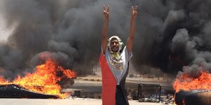 Ein Mann in sudanesischer Flagge gehüllt. Er hebt beide Arme hoch und befindet sich vor einer Rauchwand.
