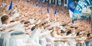 Fußballfans im Stadion tragen weiße Plastiksäcke, eine blaue Fahne von Schalke 04 wird geschwenkt