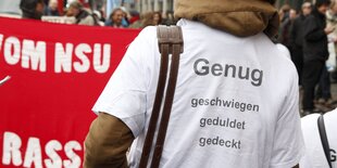 Ein Mann mit einem T-Shirt "Genug geschwiegen, geduldet, gedeckt" steht vor einem roten Demotransparent