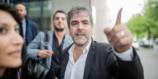 Deniz Yücel steht vor dem Berliner Amtsgericht Tiergarten, er trägt ein weißes Hemd und ein Sakko und zeigt mit dem Finder aus dem Bild.