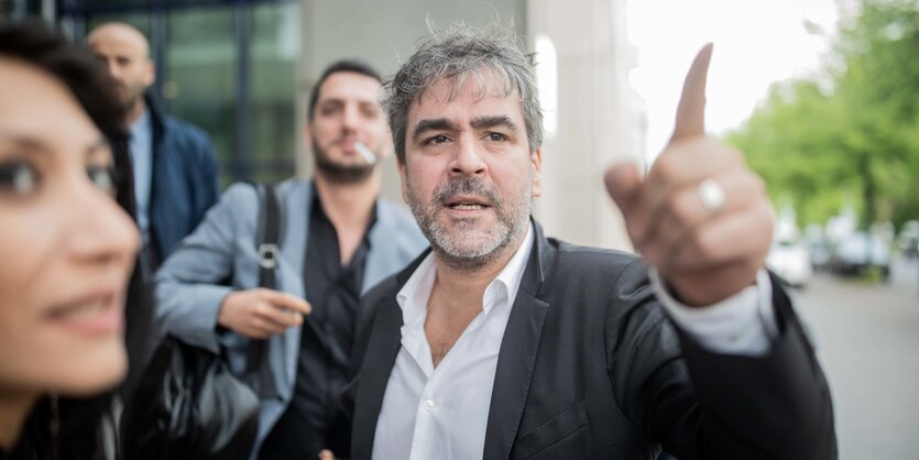 Deniz Yücel steht vor dem Berliner Amtsgericht Tiergarten, er trägt ein weißes Hemd und ein Sakko und zeigt mit dem Finder aus dem Bild.