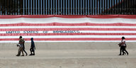 eine riesige US-Flagge hängt an einem Zaun, an dem mehrere Menschen entlang gehen