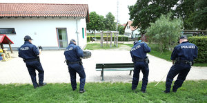 Vier Polizisten stochern mit Stöcken im Rasen vor einem öffentlichen Platz. Ansicht von hinten