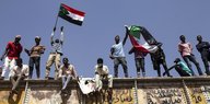 Mehrere Menschen stehen oder sitzen auf einer Mauer, einige schwenken eine sudanesische Fahne