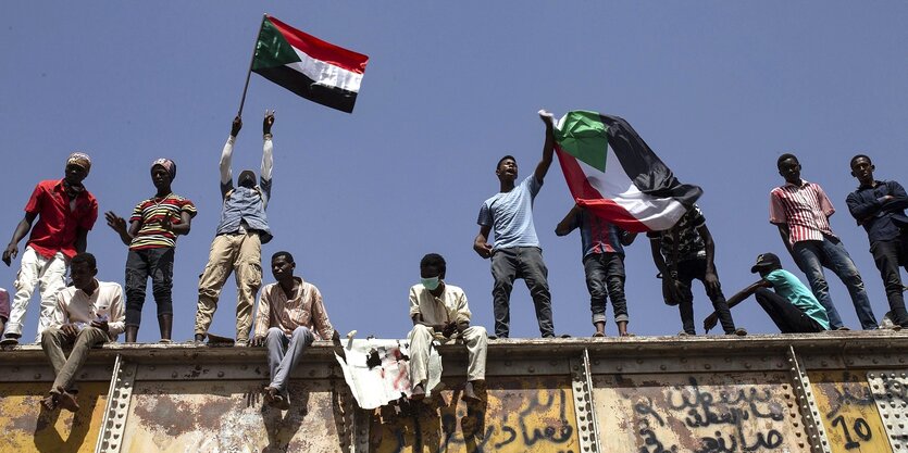 Mehrere Menschen stehen oder sitzen auf einer Mauer, einige schwenken eine sudanesische Fahne