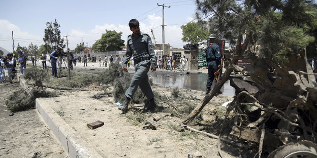 Ein afghanischer Polizist überquert eine Verkehrsinsel, neben ihm ist ein Autowrack zu sehen. Im Hintergrund sieht man mehr Polizei und Medienvertreter.