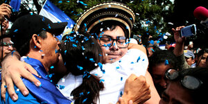 Eine Frau umarmt einen Mann, im Hintergrund eine Menschenmenge mit blau-weißen nicaraguanischen Fahnen